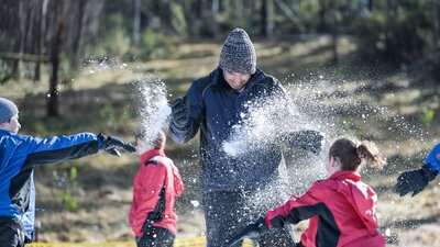 Kids throw snowballs at their dad. Snow is spraying and there is a forest in the background.