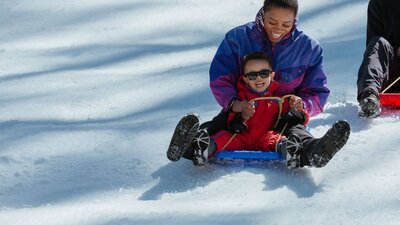 A mother and son riding a toboggan down a slope covered in snow, with big smiles on their faces.