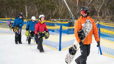 An instructor leads 3 kids, all carrying snowboards, across snow at the bottom of a ski hill